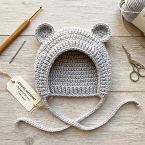May include: A light gray crochet baby bonnet featuring two small rounded bear ears on top and long chin ties. A tag attached with twine reads: HANDMADE BABY BONNET - Newborn (0-3m) - 5mm Hook. A wooden crochet hook and metal scissors are nearby.