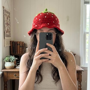Può includere: Un cappello a secchiello lavorato all'uncinetto di colore rosso a forma di fragola, caratterizzato da un picciolo verde sulla parte superiore e piccoli punti bianchi ricamati sparsi sulla superficie per imitare i semi. Tesa larga e flessibile.