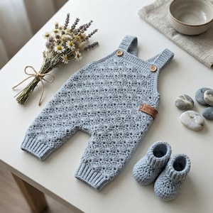 May include: A light blue crochet romper featuring a textured shell stitch pattern, two wooden buttons at the shoulder straps, and a small brown leather tag labeled handmade with love. A pair of matching light blue crochet booties are included.