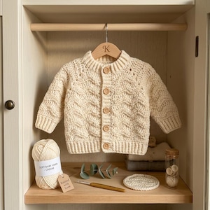 May include: A cream-coloured, long-sleeved, hand-crocheted cardigan featuring a textured cable-knit pattern, a ribbed collar, cuffs, and hem, and five light brown wooden buttons down the front, hanging on a wooden hanger with a letter K engraving.