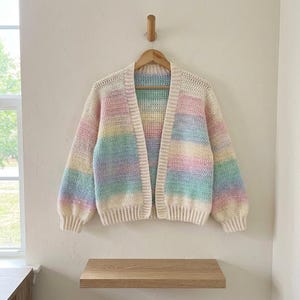 Könnte beinhalten: Eine langärmelige, vorne offene Strickjacke mit horizontalem Streifenmuster in sanften Pastellfarben wie Rosa, Gelb, Blau und Grün. Das Kleidungsstück ist in einer strukturierten Häkeloptik gefertigt und hat einen cremefarbenen Rippsaum.