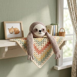 Peut inclure: Une peluche paresseux crochetée avec une tête ronde marron et de longs bras, fixée sur une couverture carrée en granny square aux couleurs pastel jaune, pêche et vert sauge. Le paresseux a un visage crème, des yeux noirs et un nez brodé.