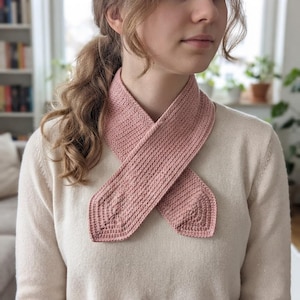 May include: A dusty rose-colored crochet scarf featuring a dense, textured stitch pattern. The scarf is designed with two pointed ends that overlap at the center, creating a layered appearance. The fabric has a consistent, uniform weave throughout.
