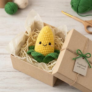 May include: A handmade crochet corn plush toy with a smiling face, nestled in a brown gift box with paper filler. The box is tied with twine and a green bow, with a tag that reads "Handmade with Love".