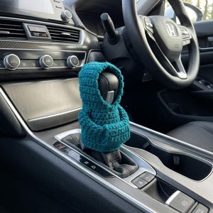 Puede incluir: Una funda de ganchillo color verde azulado para la palanca de cambios, diseñada como una sudadera con capucha en miniatura. La prenda cuenta con capucha y bolsillo frontal, tejida con punto texturizado sobre la consola central del coche.