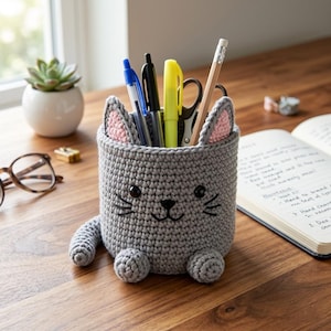 May include: A gray crocheted cat-shaped pencil holder featuring pink inner ears, black embroidered eyes, a nose, and whiskers. The cylindrical container has a small tail and two front paws at the base, holding a blue pen, black pen, yellow highlighter, scissors, and a wooden pencil.