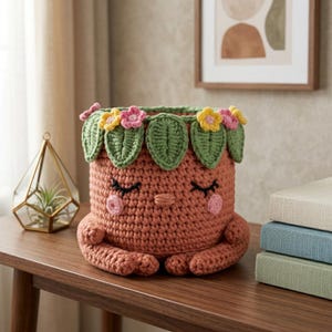 May include: A terracotta-colored crochet basket featuring a smiling face with closed eyes and pink cheeks. The top rim is decorated with a row of green crocheted leaves interspersed with small yellow and pink crocheted flowers.