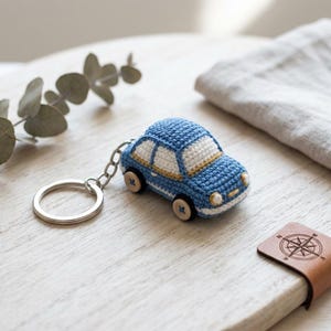 May include: A blue and white crocheted car keychain featuring a small metal ring. The car has white windows, a gold-colored trim line, and four wheels made from small, light-colored wooden buttons with blue stitching in the center.