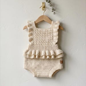 May include: A cream-coloured crochet romper featuring a textured square bodice, ruffled shoulder straps, and a double-layered ruffled waist detail. The garment is crafted with a dense stitch pattern and includes a small brown rectangular brand tag.