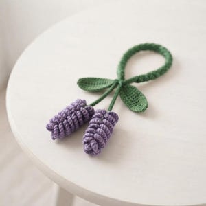 Peut inclure: Un accessoire en crochet fait main composé de deux formes de fleurs violettes texturées attachées à des tiges et des feuilles vertes, reliées à une boucle tressée verte. L'objet est réalisé en fil avec des mailles visibles sur l'ensemble.
