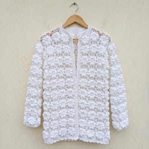 Puede incluir: Un cárdigan blanco de manga larga con un intrincado patrón de ganchillo de motivos florales en relieve por toda la prenda. Tiene un cuello redondo, un diseño abierto en la parte delantera y está colgado en una percha de madera.