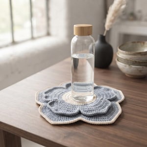 May include: A clear glass water bottle with a light brown wooden cap sits atop a flower-shaped crochet coaster. The coaster features a cream-coloured centre surrounded by layered, scalloped petals in a muted blue yarn with a cream-coloured border.