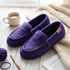 Może przedstawiać: Para ciemnofioletowych szydełkowych kapci o teksturowanym splocie, z małą brązową prostokątną metką z napisem "HANDMADE" z boku. Kapcie wykonane są z włóczki i mają klasyczny fason przypominający mokasyny z zaokrąglonym noskiem.