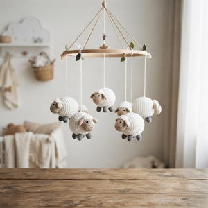 Op de afbeelding: Een hangende babymobiel met een rond houten frame opgehangen aan witte koorden. Vijf gehaakte schaapjes van crème en lichtbruin garen hangen aan het frame, elk met kleine grijze voetjes. Kleine viltblaadjes en sterretjes versieren de houten ring.