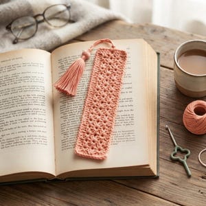 May include: A rectangular peach-coloured crochet bookmark featuring a repeating star-like pattern and a matching yarn tassel attached to the top. The bookmark is resting on the open pages of a book with visible printed text.