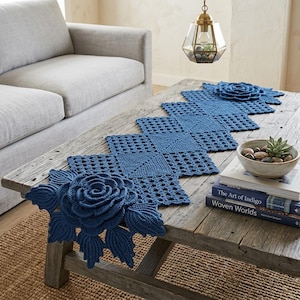 May include: A long, rectangular blue crochet table runner featuring a series of interconnected square motifs with intricate patterns. Large, three-dimensional crocheted rose blossoms with detailed leaves are attached to both ends of the runner.
