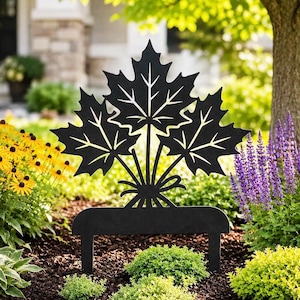 May include: A black metal garden stake featuring a cutout design of three maple leaves bundled together with a small bow at the base. The silhouette is flat and symmetrical, with pointed leaf edges and detailed vein cutouts throughout the foliage.