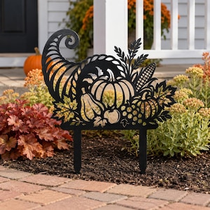 Puede incluir: Un adorno de jardín de metal negro con un diseño recortado de un cuerno de la abundancia lleno de calabazas, calabacines, uvas, espigas de trigo y hojas de otoño. La silueta se sostiene mediante dos finas varillas de metal para el suelo.