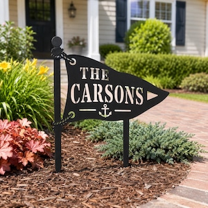 Puede incluir: Un letrero de jardín de metal negro con forma de banderín sujeto a un poste vertical. El letrero presenta el texto THE CARSONS en letras recortadas sobre un pequeño símbolo de ancla. El diseño incluye detalles que imitan una cuerda.