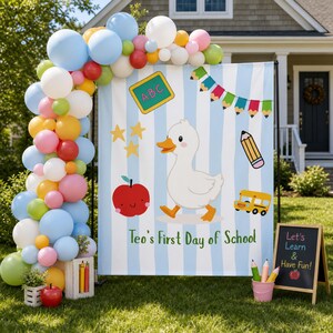 Peut inclure: Une toile de fond avec un canard blanc, une pomme rouge, un bus scolaire jaune et un tableau noir avec le texte Teo's First Day of School, entourée d'une arche de ballons bleus, blancs, roses, verts et jaunes, avec un petit tableau noir.
