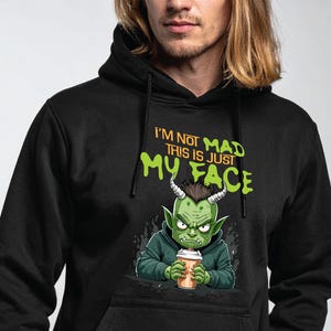 Puede incluir: Sudadera con capucha negra con un diseño de un duende de dibujos animados sosteniendo una taza de café. El duende verde tiene cuernos y una expresión seria. El texto dice "I'M NOT MAD THIS IS JUST MY FACE".