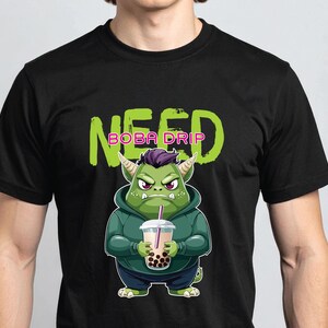 May include: Black t-shirt with a green monster graphic holding a boba drink. The monster has purple hair, green skin, and is wearing a green hoodie. The text "NEED BOBA DRIP" is above the graphic in green and pink.