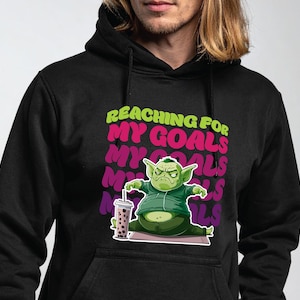 Può includere: Felpa con cappuccio nera con un personaggio dei cartoni animati verde che tiene un bubble tea. Il testo "REACHING FOR MY GOALS" è sopra il personaggio in verde e rosa.