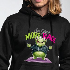 Pode incluir: Moletom preto com um monstro verde de desenho animado em pose de ioga em um tapete roxo. O monstro tem olhos vermelhos e chifres. O moletom tem o texto "Less Yoga, More Rage" em verde e rosa.