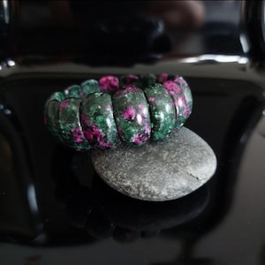 Può includere: Un braccialetto di perline di pietre verdi e rosa lucide poggia su una pietra grigia. Le perline del braccialetto sono un mix di verde scuro e rosa vibrante, creando un contrasto sorprendente. Lo sfondo è una superficie nera lucida.