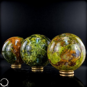 Peut inclure: Trois pierres sphériques polies présentant un mélange vibrant de tons terreux vert mousse, olive et brun profond. Chaque sphère minérale lisse et arrondie est présentée sur un petit support métallique circulaire de couleur dorée sur fond sombre.