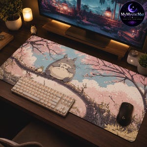 May include: A rectangular desk mat featuring a printed illustration of a grey, round creature sitting on a tree branch amidst pink cherry blossoms and a light blue sky. A white mechanical keyboard and a black computer mouse rest on the mat.