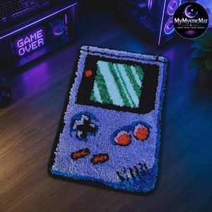Op de afbeelding: Een paars en zwart tapijt in de vorm van een retro handheld gameconsole. Het tapijt heeft een groen en wit scherm, oranje knoppen en een navigatiepad. De tekst "Dip-ite" is zichtbaar. De tekst "GAME OVER" wordt weergegeven op een neonbord.
