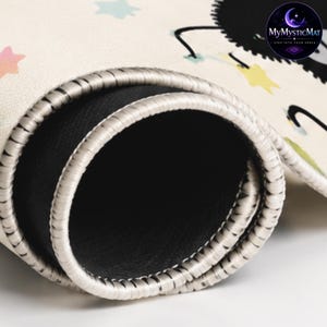 May include: A rolled-up cream-colored mat with a black circular logo reading MyMysticMat, Step Into Your Space. The mat features a pattern of small pastel-colored stars and a black character. It has a black rubberized underside and stitched edges.