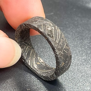 Peut inclure: Une bague gris foncé présentant un motif de Widmanstätten gravé distinct sur toute sa surface. L'anneau a un profil plat et une texture métallique mate, mettant en valeur des lignes géométriques entrecroisées caractéristiques des météorites.