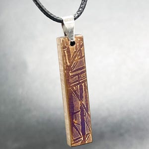 Pode incluir: Um pendente retangular com um padrão geométrico castanho e roxo com acabamento metálico. O pendente está suspenso por um cordão preto através de um passador de metal prateado, criando uma orientação vertical para esta peça de joalharia.