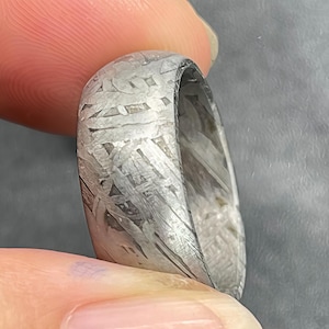 Puede incluir: Un anillo de tono plateado que presenta un patrón de Widmanstätten distintivo en su superficie. La banda de metal muestra una estructura cristalina compleja y entrelazada con grabados lineales en gris claro y oscuro por todo el exterior.