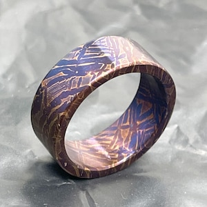 Puede incluir: Un anillo con un patrón de Widmanstätten distintivo y acabado metálico. La superficie muestra una red compleja y entrecruzada de líneas de color azul profundo y cobre, creando una textura geométrica y cristalina en toda la banda.