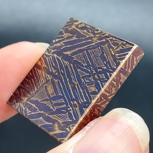 Puede incluir: Una pieza rectangular de meteorito Muonionalusta con un patrón de Widmanstätten distintivo. La superficie muestra un contraste metálico entre áreas grabadas en tono azul oscuro y líneas cristalinas de color marrón dorado claro.