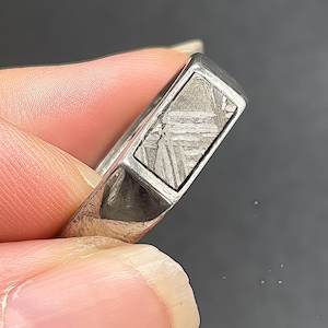 Könnte beinhalten: Ein silberfarbener Ring mit einer rechteckigen Einlage, die ein ausgeprägtes Widmanstätten-Gefüge aufweist. Die Metallschiene hat eine polierte Oberfläche mit sichtbaren texturierten Details an den Seiten und einem geometrischen Design.