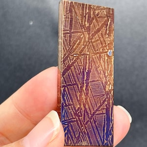 May include: A rectangular metallic piece featuring a distinct Widmanstätten pattern with intersecting lines in shades of bronze, copper, and deep violet. The surface displays a polished, etched texture with a visible crystalline structure throughout.