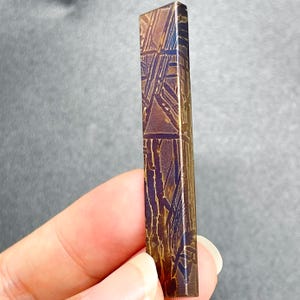 May include: A long, rectangular meteorite fragment featuring a distinct Widmanstätten pattern of intersecting metallic lines. The surface displays a metallic bronze and dark brown finish with a geometric grid of etched lines across its length.
