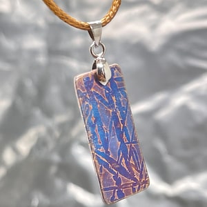 May include: A rectangular pendant featuring a metallic surface with a blue and copper-toned abstract pattern. The pendant is suspended from a silver-colored metal bail attached to a thin, light brown cord, creating a simple necklace design.