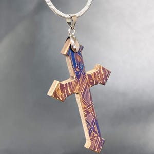 May include: A cross pendant featuring a layered wood construction with a distinct purple and brown geometric pattern on the front surface. The pendant is attached to a silver-toned metal bail and hangs from a matching silver-toned snake chain necklace.