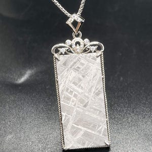 May include: A rectangular pendant featuring a metallic grey meteorite slice with a distinct Widmanstätten pattern. The piece is framed in silver-toned metal with twisted rope edges and topped with a decorative filigree bail set with small clear stones.
