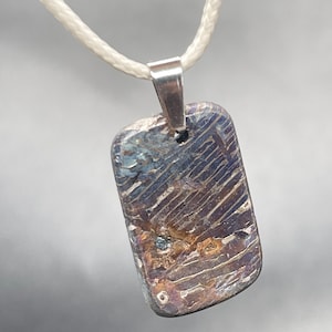 May include: A rectangular pendant with a metallic, iridescent surface featuring deep blue, purple, and copper-toned streaks and a textured, grooved pattern. The pendant is attached to a silver-toned metal bail and hangs from a thin, off-white cord.