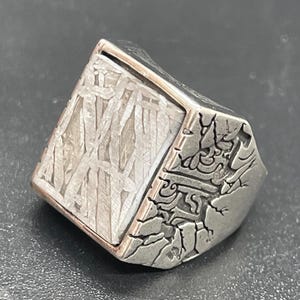 Peut inclure: Une bague argentée dotée d'une face rectangulaire avec un motif métallique entrecroisé. Les côtés de la bague présentent un aspect sombre, texturé et fissuré, contrastant avec l'incrustation géométrique plus claire sur le dessus.