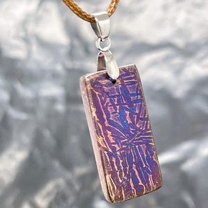 May include: A rectangular pendant featuring a metallic, iridescent surface with a dark purple and blue cross-hatched pattern. The pendant is attached to a silver-toned metal bail and hangs from a braided brown cord necklace.