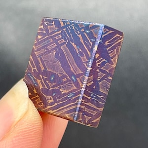May include: A rectangular piece of etched meteorite featuring a distinct Widmanstätten pattern with deep purple and bronze metallic tones. The surface displays a complex, intersecting crystalline structure with sharp geometric lines throughout.