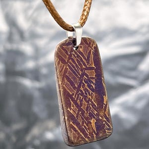 May include: A rectangular pendant featuring a metallic surface with a dark purple and bronze-toned Widmanstätten pattern. The pendant is suspended from a brown braided cord by a silver-colored metal bail, showcasing a distinct crystalline structure.