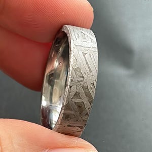 May include: A silver-toned metal ring featuring a distinct, intricate Widmanstätten pattern across its outer surface. The band has a flat profile with a polished, reflective interior surface that contrasts with the textured, etched exterior finish.